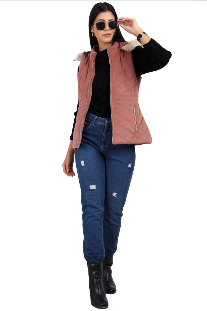 A woman in a standing pose wearing shades, coatsnmore’s half sleeves jacket in onion pink colour with a textured pattern, a removable hood with faux fur trim, side zippered pockets and a zip closure and blue jeans, holding her shades from her left hand.